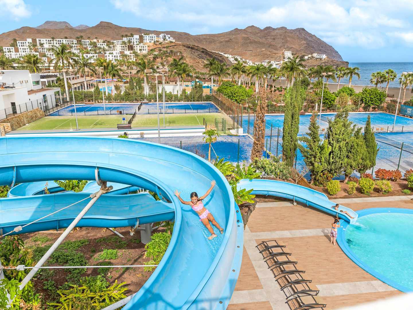 blte6a583a3e8ba97c6-spanien-kanaren-fuerteventura-playitas-aparthotel-h-DER1516558.jpg Kind rutscht auf blauer Wasserrutsche im Playitas Aparthotel mit Blick auf Tennisplätze und das Meer
