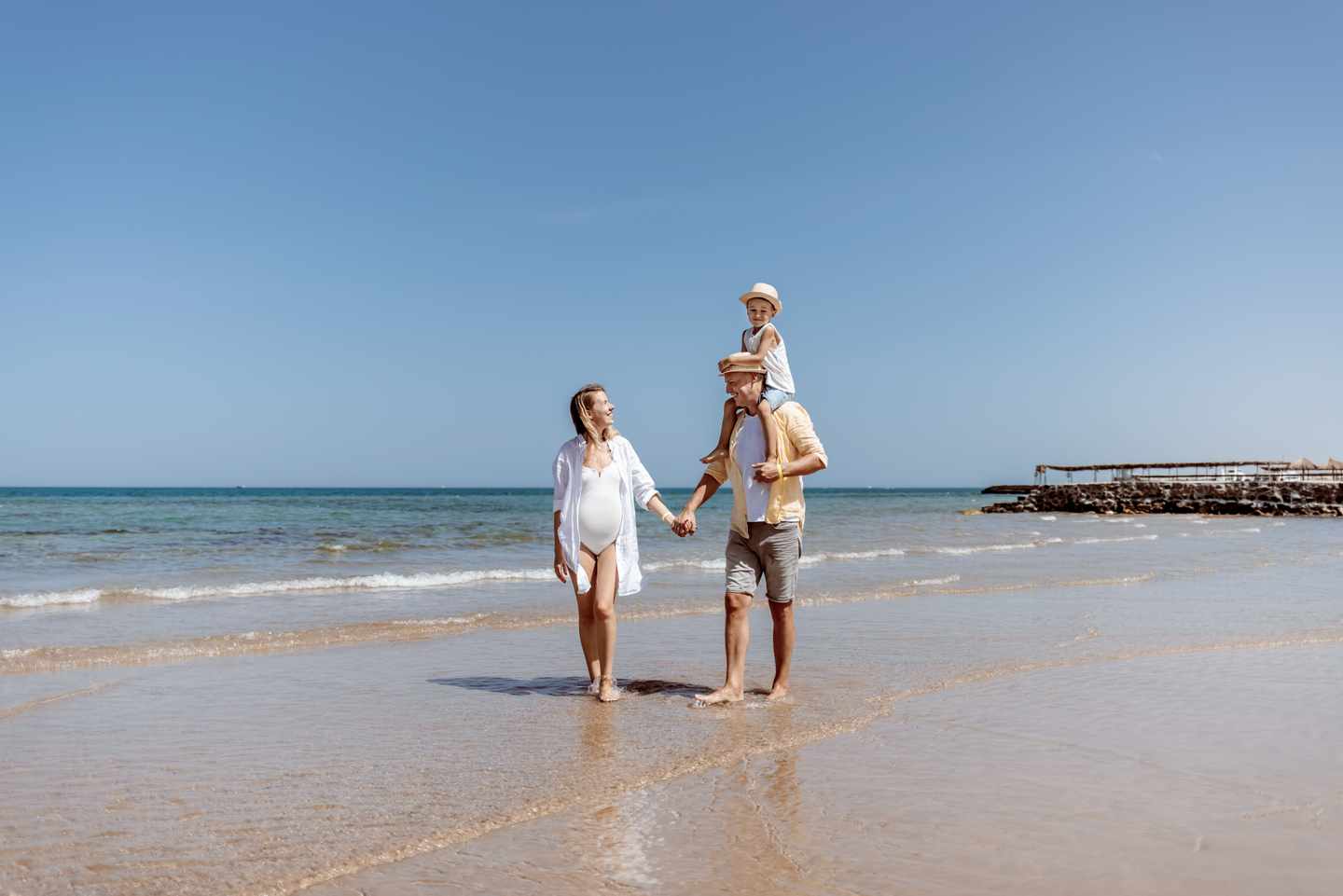 Familie mit Kind spaziert barfuß am flach abfallenden Sandstrand von Hammamet entlang der Mittelmeerküste