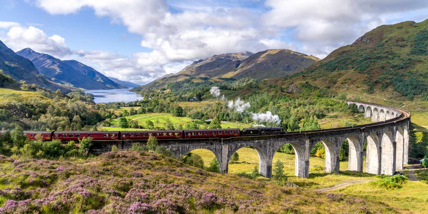 Ein Dampfzug fährt über die Bögen des Glenfinnan-Viadukts durch ein grünes Tal in den schottischen Highlands