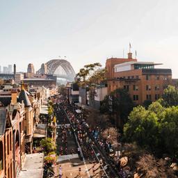 Blick auf Straße in Sydney beim Marathon mit Läufern und im Hintergrund ist die Harbour Bridge und das Opernhaus in weiß zu sehen.