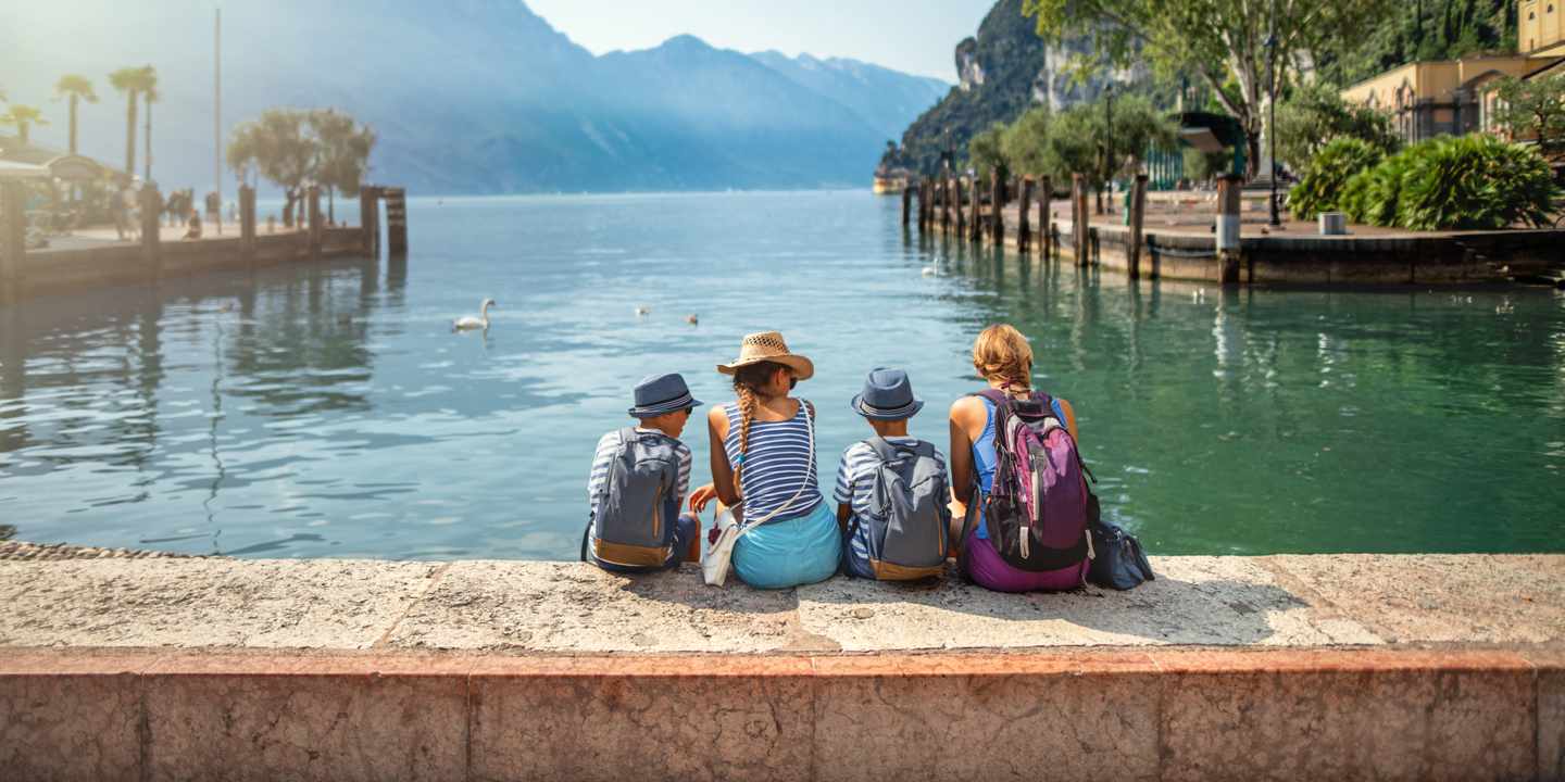 Zwei Erwachsene und zwei Kinder sitzen mit dem Rücken zur Kamera am Ufer eines Sees, im Hintergrund Berge und ein Schwan im Wasser.