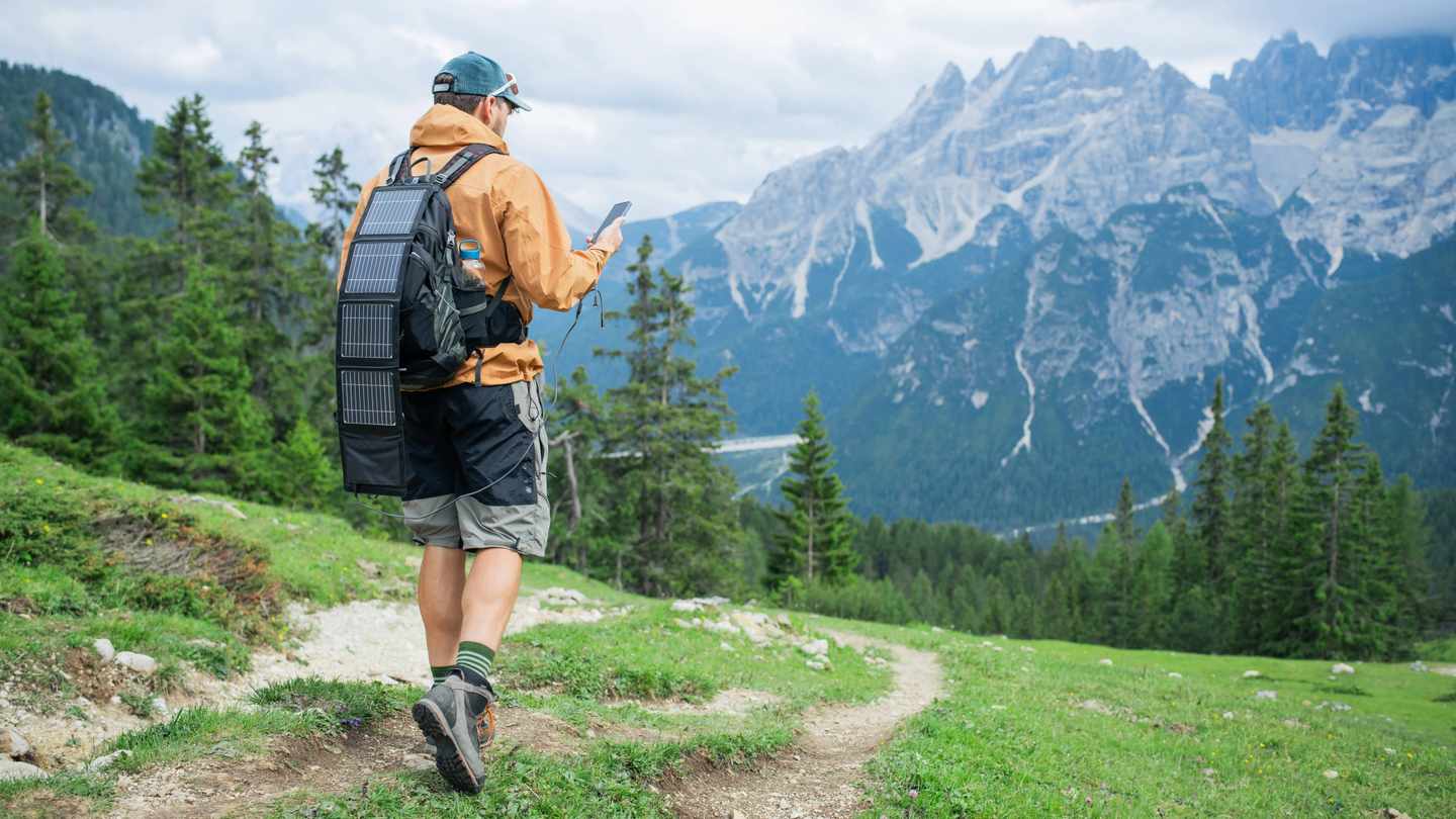 Mann mit tragbarem Solarpanel auf dem Rucksack lädt Smartphone beim Wandern in den Alpen nachhaltig in der Natur auf