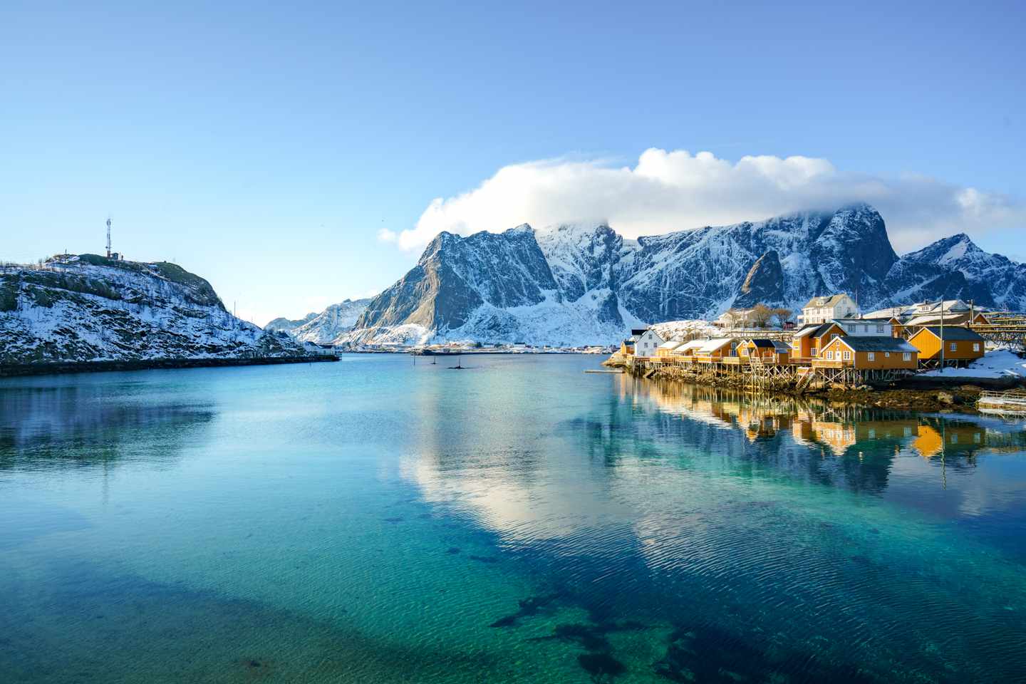 Bucht auf den Lofoten mit ruhigem Wasser, schneebedeckten Bergen und traditionellen Holzhäusern am Ufer