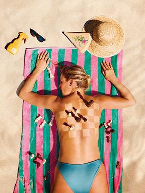 Angebote entdecken Last Minute 2026: Frau liegt auf einem Strandtuch im Sand, auf ihrem Rücken ist ein Schachbrett mit Figuren. Neben ihr liegen Sonnencreme, Hut, Brille und Fächer