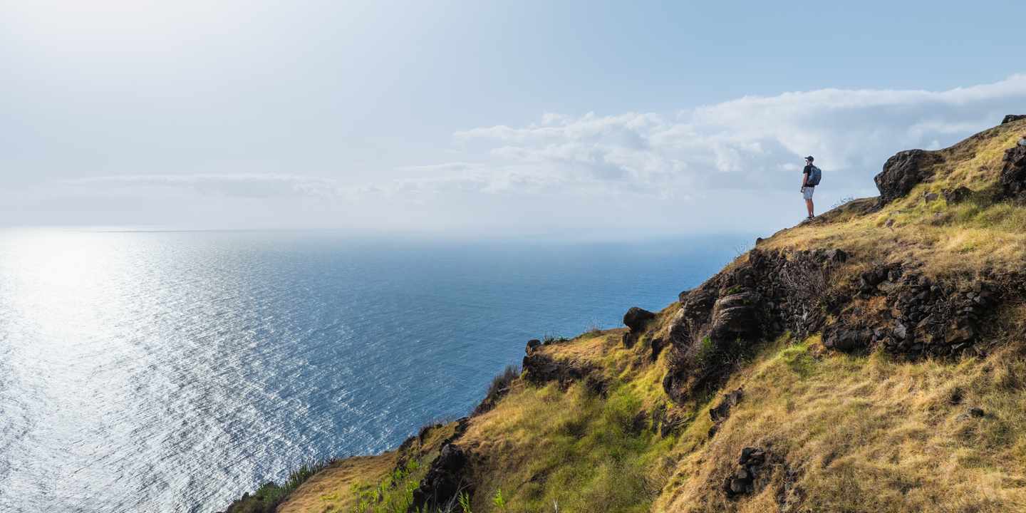 Wanderer steht an einer grün bewachsenen Klippe und schaut auf den blauen Ozean