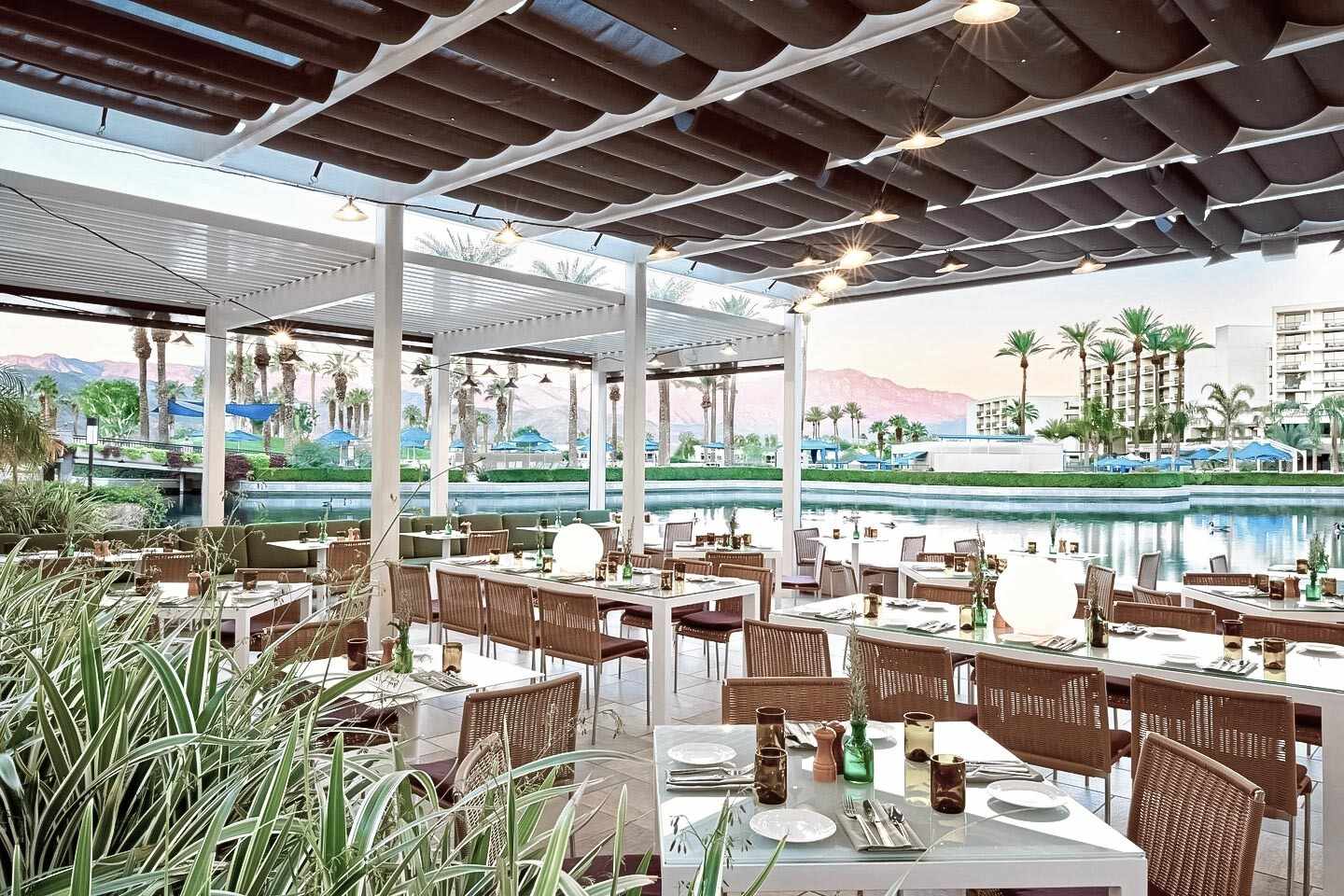 Überdachte Außenterrasse des Coachella-Hotels JW Marriott Desert Springs Resort & Spa mit gedeckten Tischen, im Hintergrund der große Pool mit Palmen und die Berge