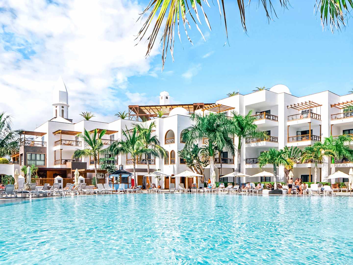 blt8bfabeab0863e5cb-spanien-kanaren-lanzarote-playa-blanca-pool-hotelanlange-h-princesa-yaiza-suite-hotel-resort-DER1669525.jpg Poolbereich des Hotels mit dahinterliegendem weißen Gebäude und grünen Palmen