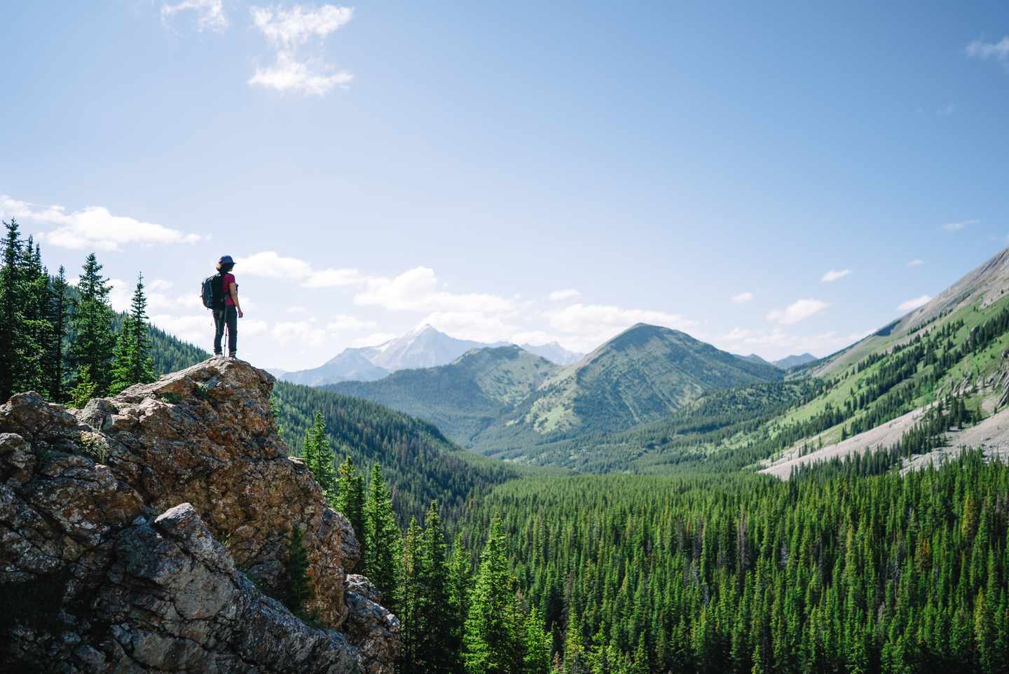 Alleinreisende Wanderin mit Rucksack steht auf einem Felsen und blickt über bewaldete Täler und hohe Berge