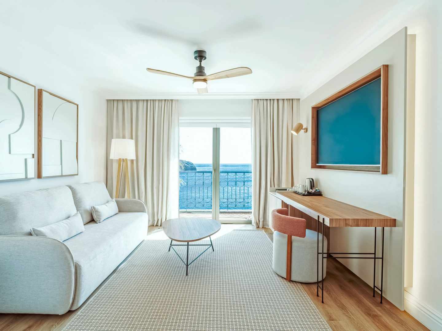 DER1588353.jpg Helles Hotelzimmer mit Sofa runder Holz-Couchtisch Schreibtisch mit Stuhl und großem Fenster mit Blick auf das Meer