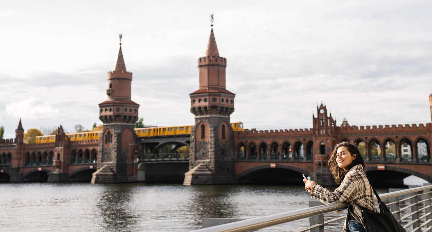 Alleinreisende Frau mit Smartphone an der Spree vor der Oberbaumbrücke und einer gelben U-Bahn in Berlin