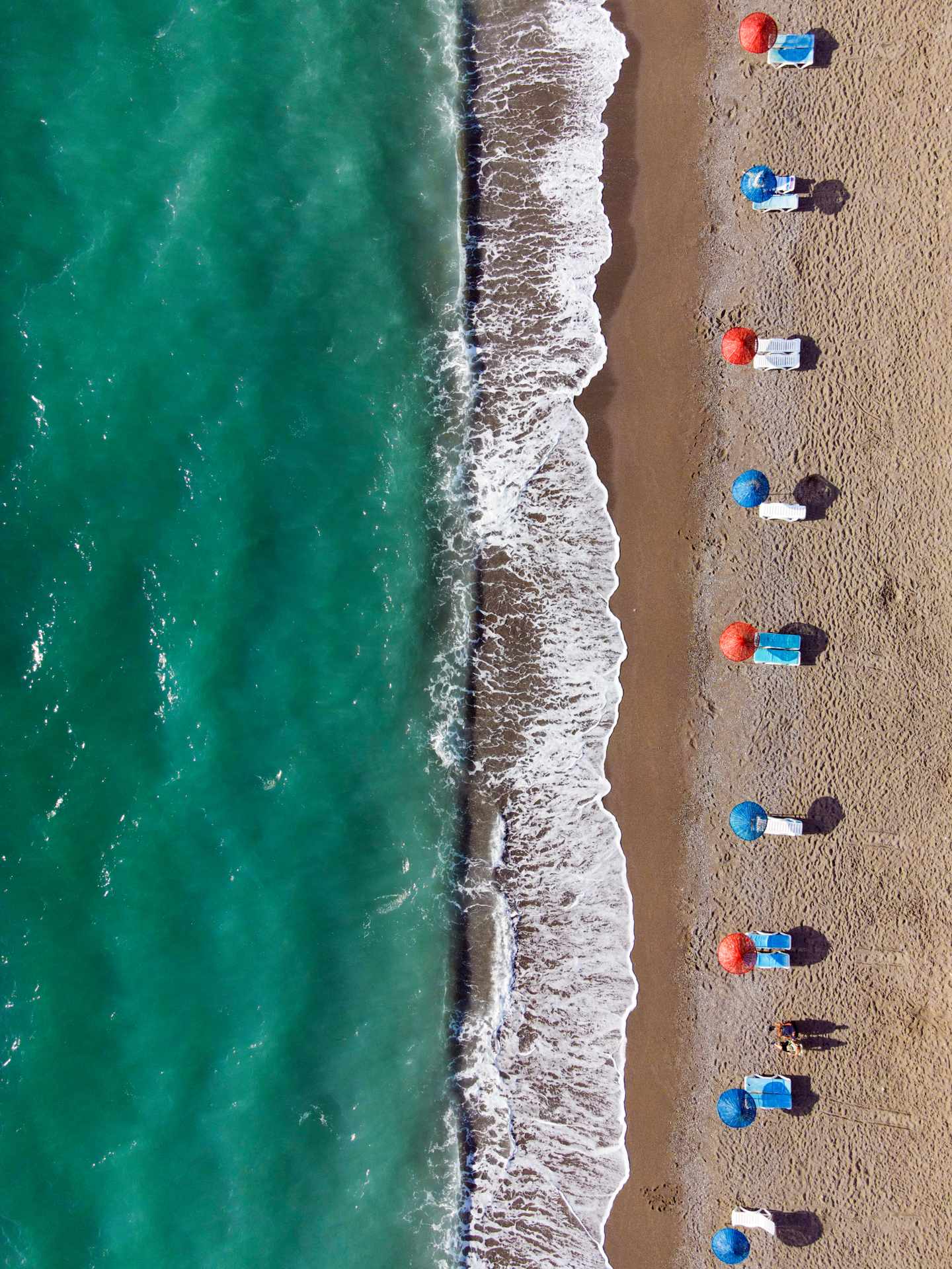 GettyImages-2253649415.jpg GettyImages-2253649415.jpg Vogelperspektive auf Sonnenschirme und Liegestühle am Strand bei Wellengang mit Brandung