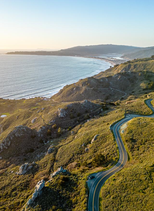 Jetzt entdecken Rundreisen: eine kurvige Straße schlängelt sich durch eine grüne Küstenlandschaft in Kalifornien, USA