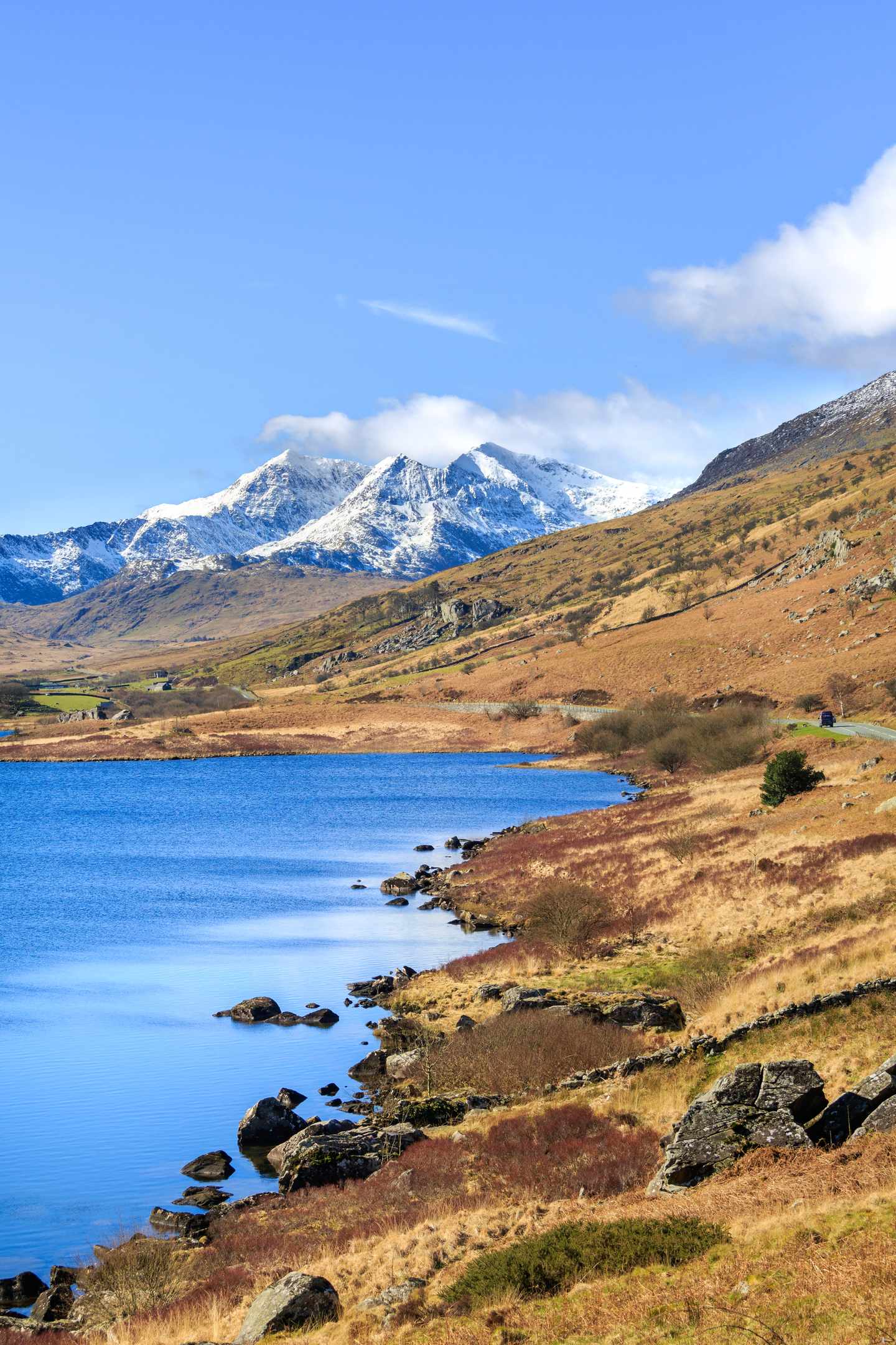 Seeufer im Snowdonia National Park mit schneebedeckten Bergen Felsen und Heideflächen unter blauem Himmel