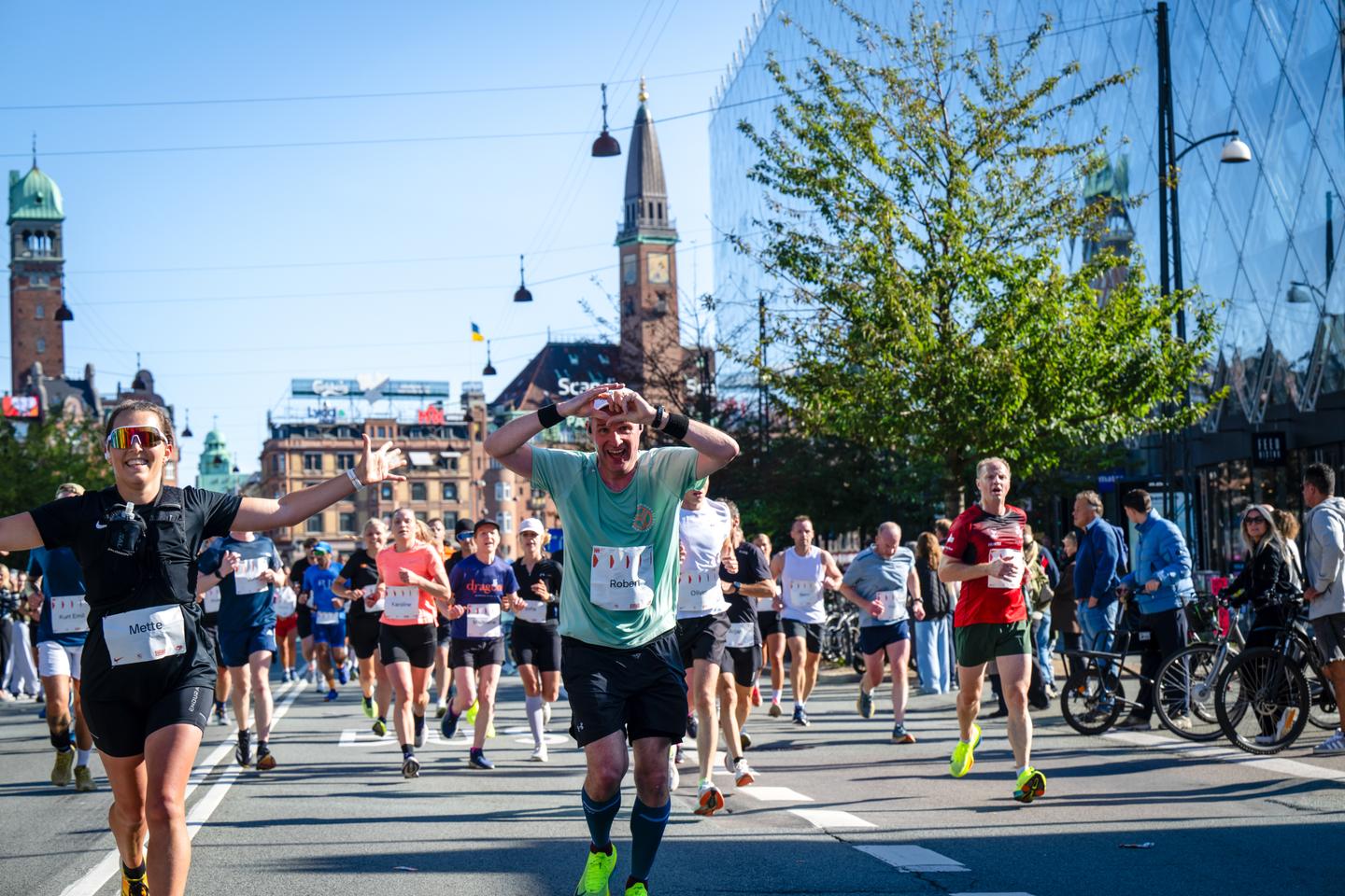Bunt gekleidete Teilnehmer des Kopenhagen Marathons laufen auf einer Straße vor einem roten Gebäude mit einem spitzen Turm und rechts ein Gebäude mit blau spiegelnder Glasfassade und Publikum davor.