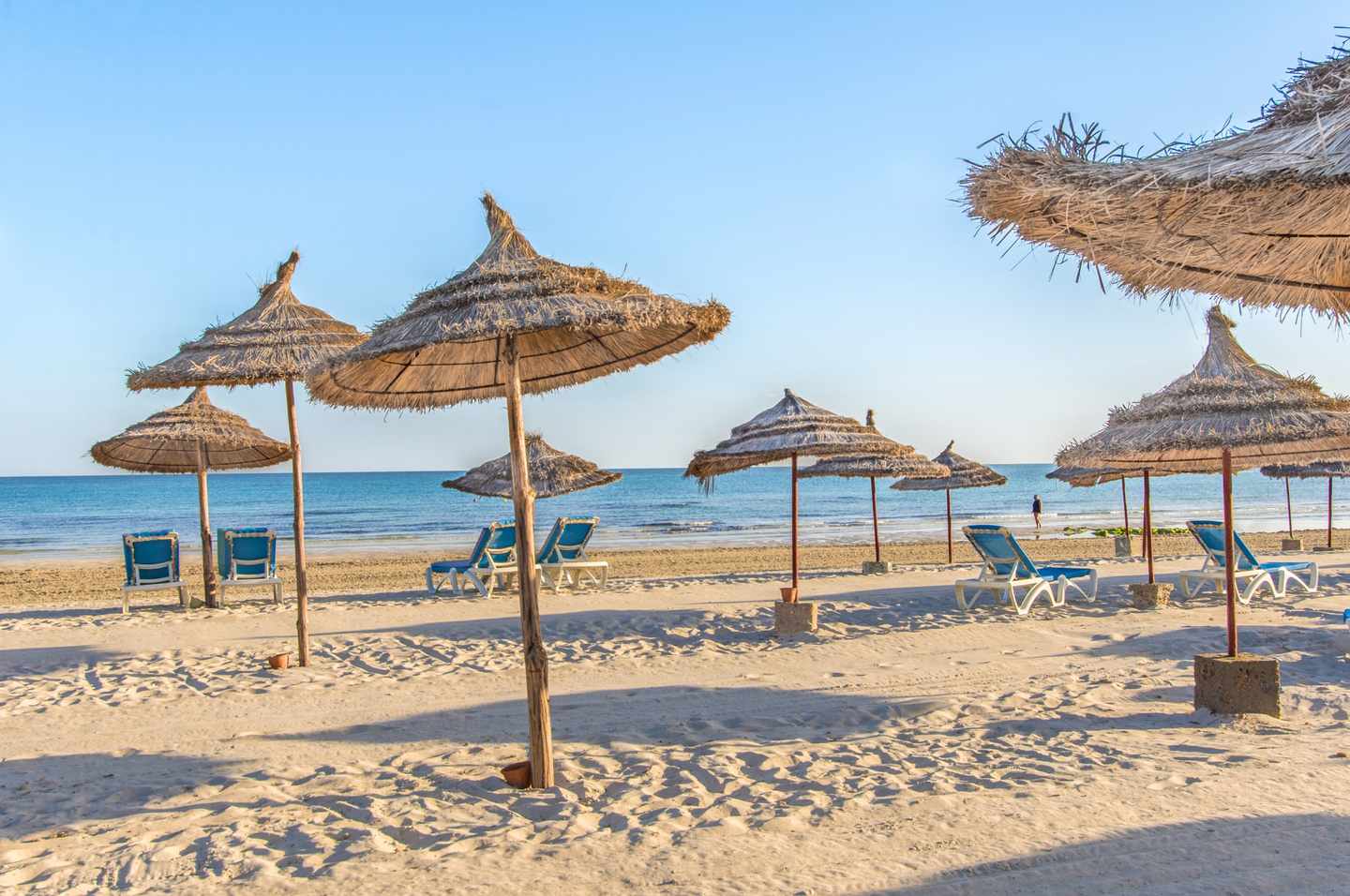 Strohschirme und Liegen am breiten Sandstrand an der Ostküste von Djerba unter klarem Himmel in Tunesien