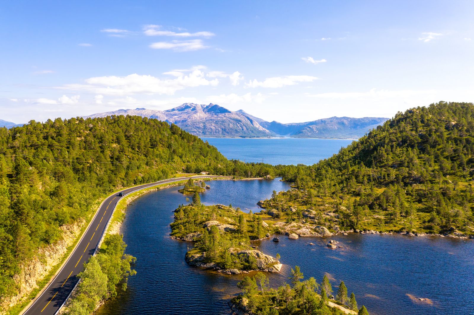 Luftaufnahme einer kurvigen Straße entlang eines Sees mit bewaldeten Hügeln und Bergen im Hintergrund
