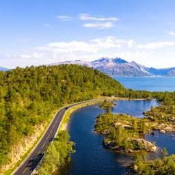 Luftaufnahme einer kurvigen Straße entlang eines Sees mit bewaldeten Hügeln und Bergen im Hintergrund