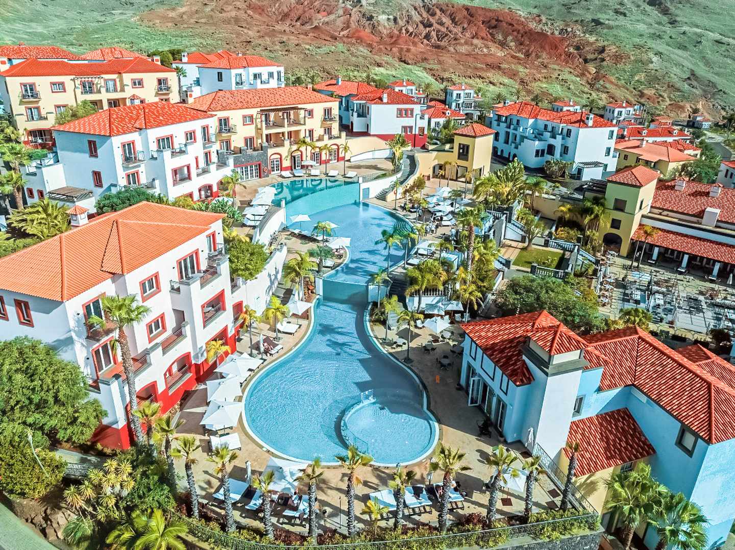 Luftaufnahme des Hyatt Ziva Madeira in Caniçal mit geschwungenem Pool und weiß-roten Gebäuden vor der Vulkanlandschaft