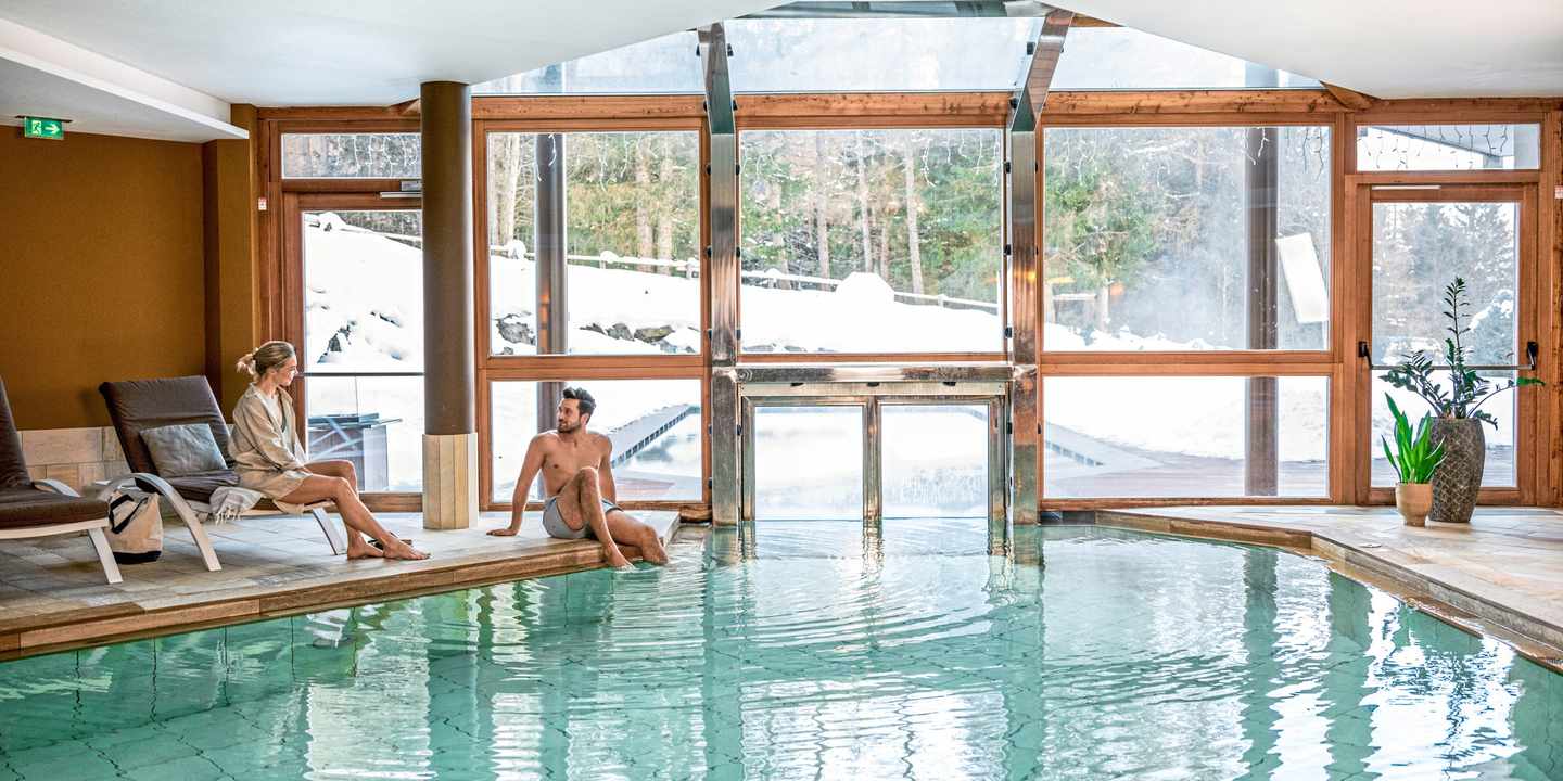 Ein Paar entspannt am Innenpool eines Hotels mit Blick auf die verschneite Berglandschaft