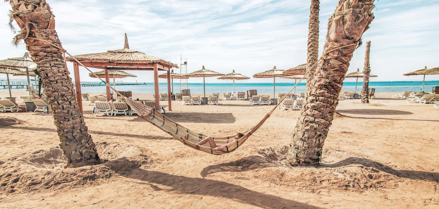 Hängematte zwischen zwei Palmen an einem Sandstrand mit Sonnenschirmen und türkisblauem Meer in Tunesien