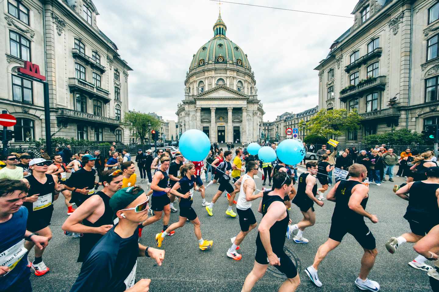 55262942862_cc3393477e_o.jpg Viele Läufer joggen in schwarzer Trainingskleidung auf einer Straße beim Kopenhagen-Marathon und ein paar von ihnen halten blaue Luftballons in der Hand.