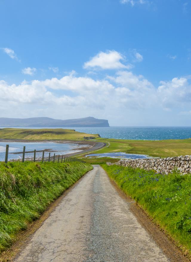 Jetzt entdecken Landschaft mit schmaler Straße zwischen grünen Wiesen Steinmauer rechts Zaun links und Blick auf Meer und bewölkten Himmel